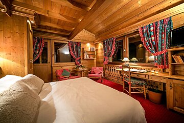 A cozy, rustic wood-paneled bedroom features a white bed, red carpet, and colorful curtains. It has a desk, armchairs, and warm lighting.