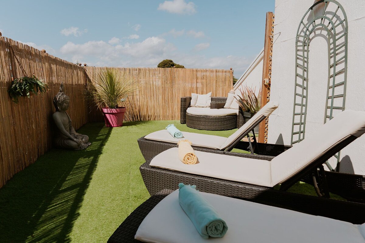 A sunny outdoor terrace features artificial turf, rattan sun loungers, a Buddha statue, and a round daybed, framed by a bamboo privacy fence.