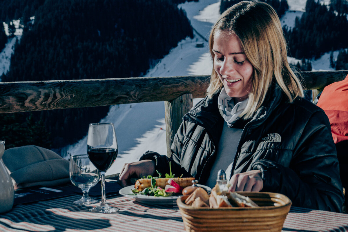 A woman wearing a north face jacket is sitting at a table with a plate of food and a glass of wine