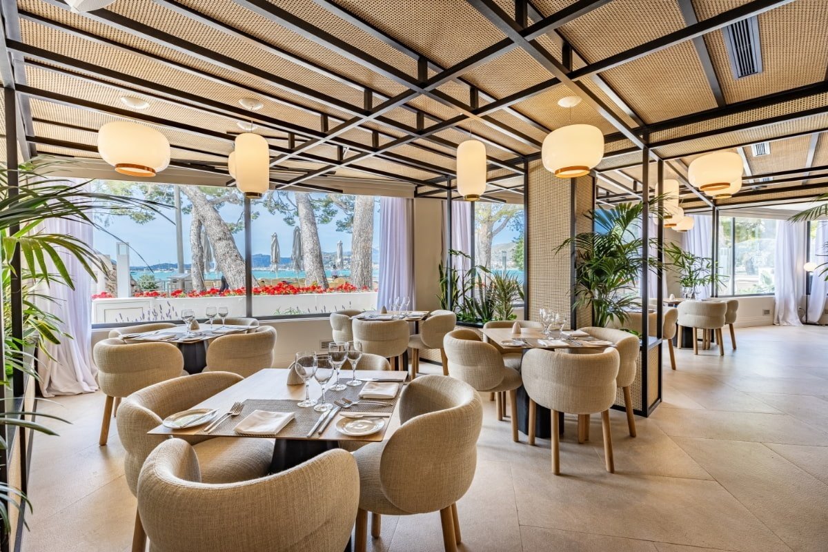 Modern restaurant with a woven ceiling, pendant lights, and large windows showcasing a beautiful sea and beach view.