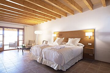 A bright, rustic-modern bedroom featuring twin beds, exposed wooden ceiling beams, tiled floors, and a patio view through sliding doors.