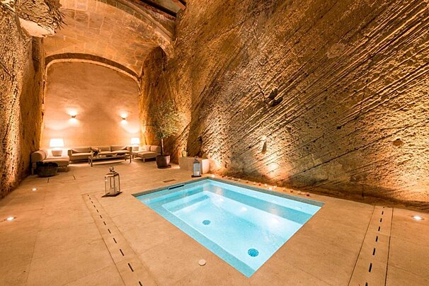 A luxurious, warm-lit underground room with a small pool, rough stone walls, an arched ceiling, and a cozy seating area.