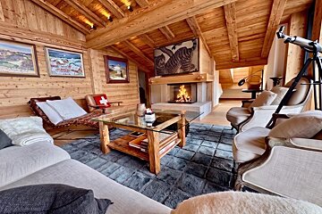Cozy, rustic cabin living room with wooden walls and ceiling, a burning fireplace, plush seating, and a glass-top coffee table.
