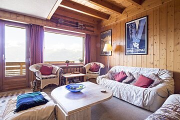 A cozy wooden cabin living room with sofas, armchairs, a coffee table, and large windows overlooking mountains.