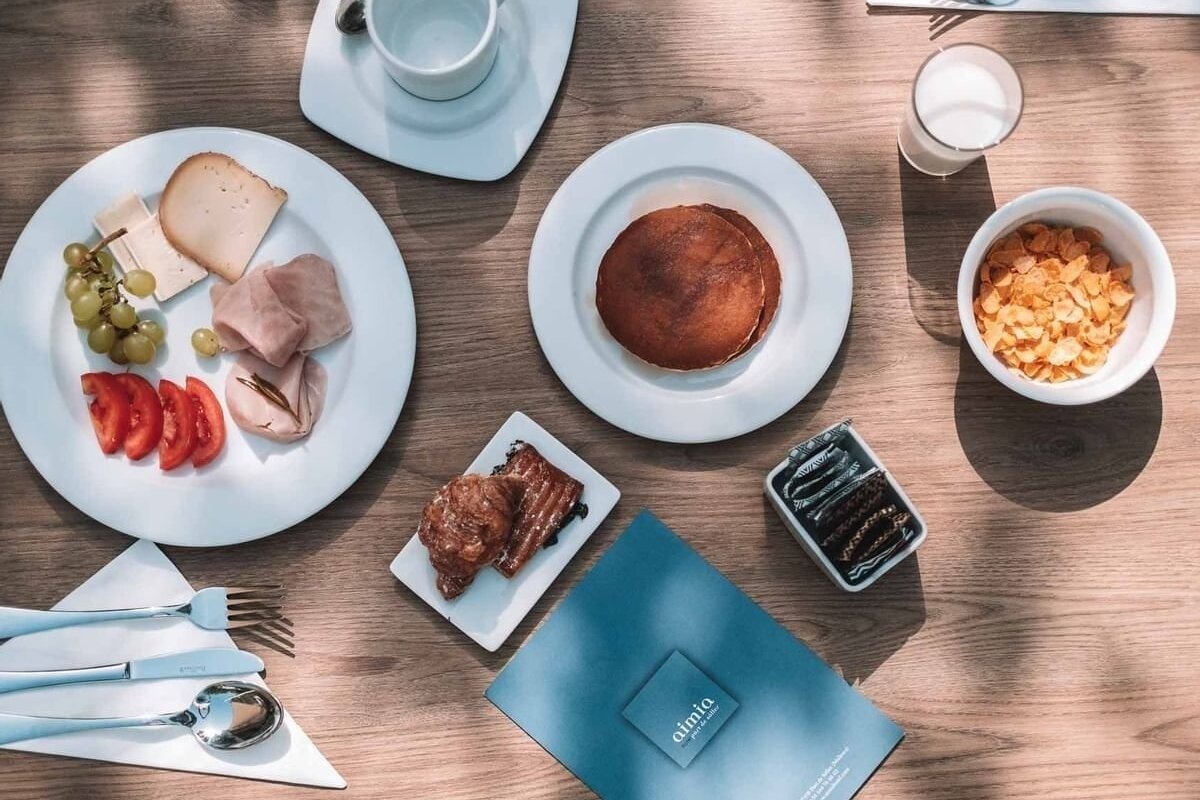 An overhead view of a diverse breakfast spread on a wooden table, featuring pancakes, cereal, cheese, ham, fruit, croissants, and milk.