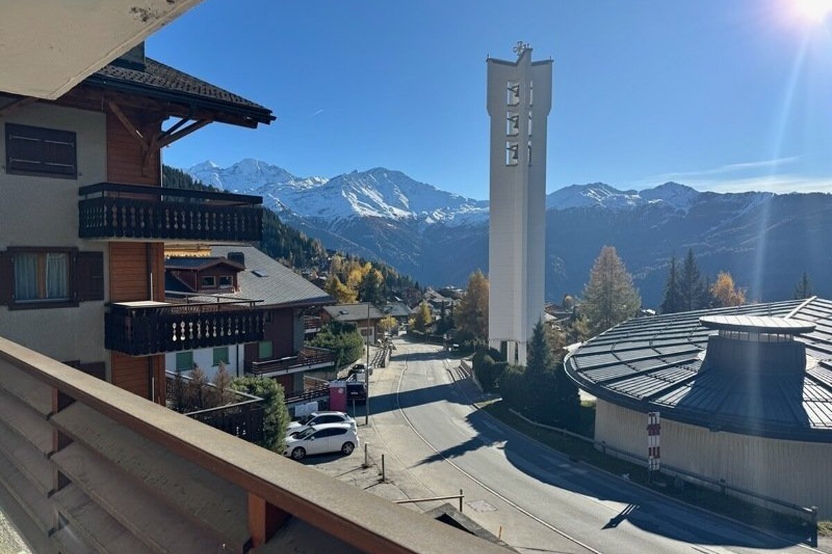 Scenic view from a balcony of an alpine village with a tall modern church tower, snow-capped mountains, and a clear blue sky.