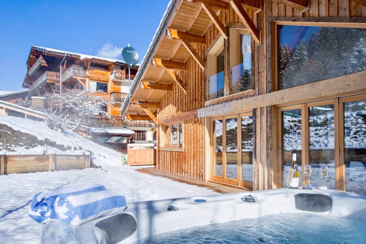 A luxurious wooden chalet in a snowy mountain landscape, featuring an inviting outdoor hot tub with champagne, fresh towels, and clear blue skies.