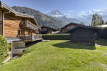 Charming wooden chalets with green lawns and balconies, set against a backdrop of towering snow-capped mountains under a clear blue sky.