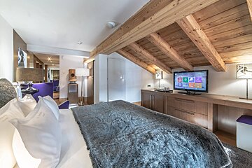 A stylish attic bedroom with exposed wooden beams, a plush bed with a grey furry blanket, a TV, and subtle purple accents.