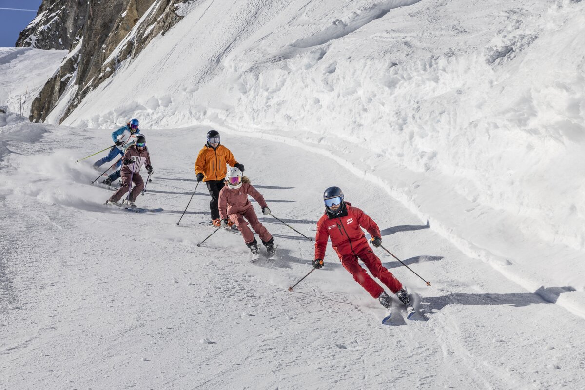 Five skiers descend a sunny, snow-covered mountain slope, kicking up powder as they carve turns with a rugged peak in the background.