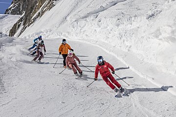 Five skiers descend a sunny, snow-covered mountain slope, kicking up powder as they carve turns with a rugged peak in the background.