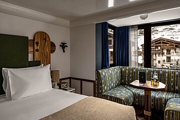 Cozy hotel room with a white bed, a vibrant green and blue patterned sofa, and large windows revealing a snowy mountain village view.
