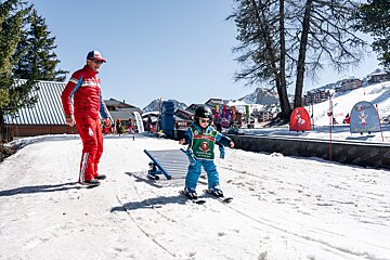 An adult in a red ski suit watches a young child learning to ski on a sunny day at a snowy ski resort with mountains in the background.