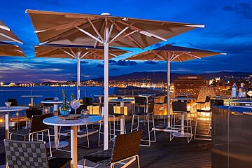A stylish rooftop bar at dusk, with umbrellas, tables, and drinks overlooking a scenic bay, city lights, and distant mountains.