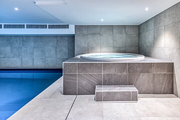 A modern indoor spa area featuring a bright blue swimming pool on the left and a tiled, built-in hot tub on the right, surrounded by gray tiles.