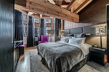 A luxurious, cozy bedroom with exposed wooden beams, a plush grey bed, dark curtains, and a vibrant purple accent chair.