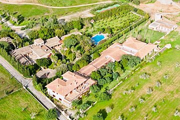 Aerial view of a sprawling estate featuring multiple terracotta-roofed buildings, lush gardens, a blue pool, tennis court, and green farmlands.