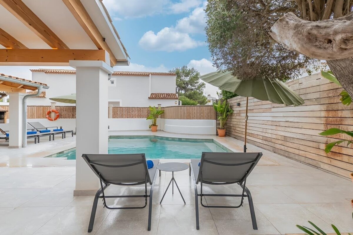 A sunny, inviting poolside retreat with two lounge chairs, a clear pool, white buildings, and lush greenery under a blue sky.