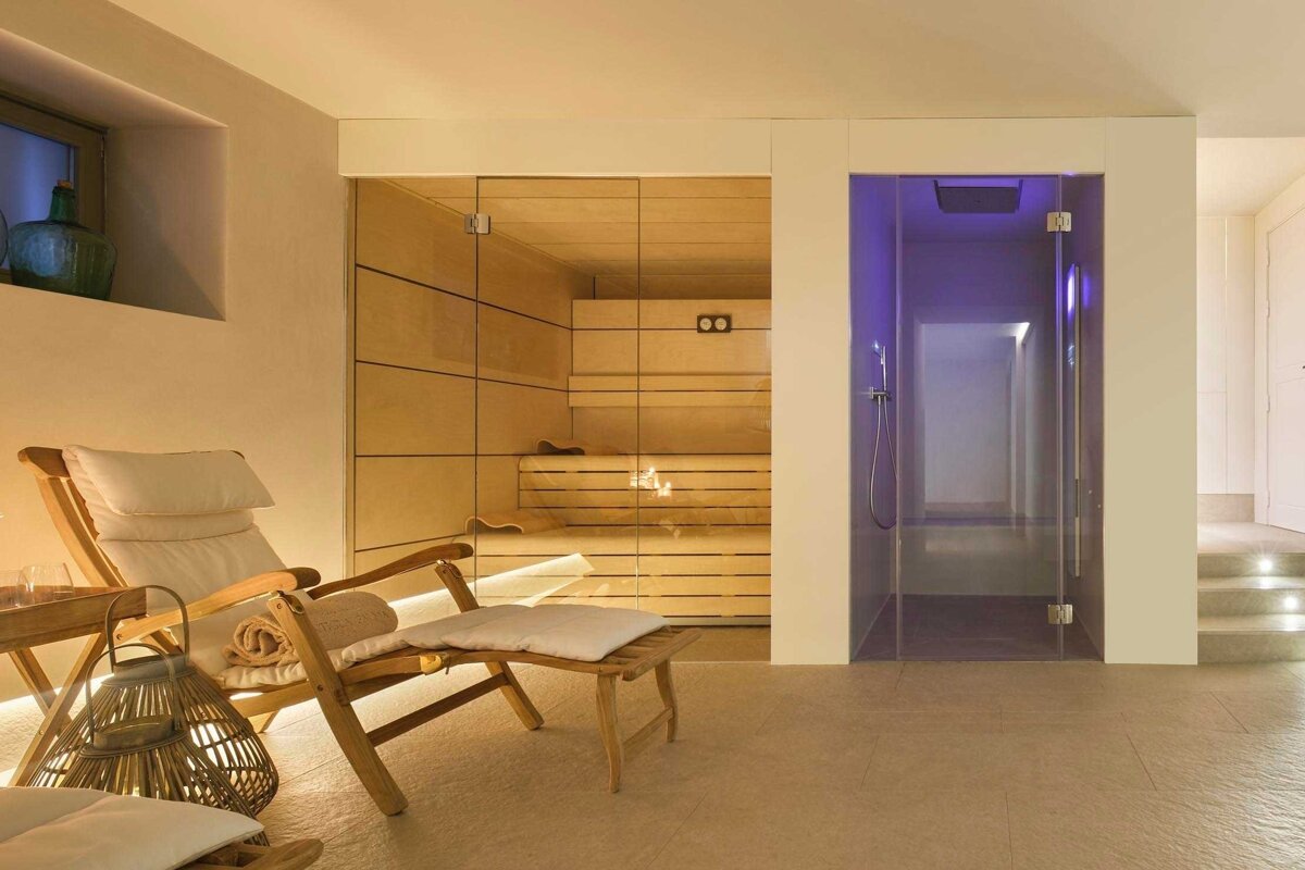 A serene spa room featuring a wooden sauna behind a glass door, a purple-lit glass shower, and two wooden loungers, all bathed in warm light.