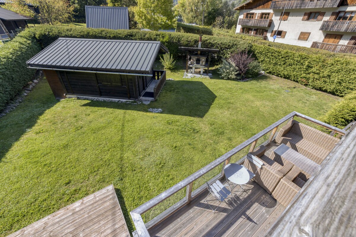 Elevated view of a green backyard with a wooden deck, outdoor furniture, a small shed, and a brick oven, surrounded by hedges and distant houses.