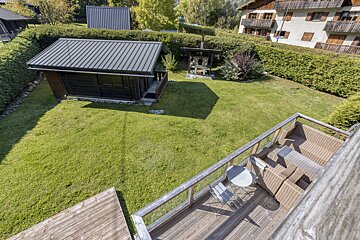 Elevated view of a green backyard with a wooden deck, outdoor furniture, a small shed, and a brick oven, surrounded by hedges and distant houses.