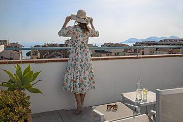 A woman in a floral dress stands on a balcony overlooking the ocean