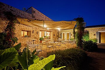 A cozy stone courtyard patio illuminated by string lights under an awning at dusk, featuring many white tables and chairs for outdoor dining.