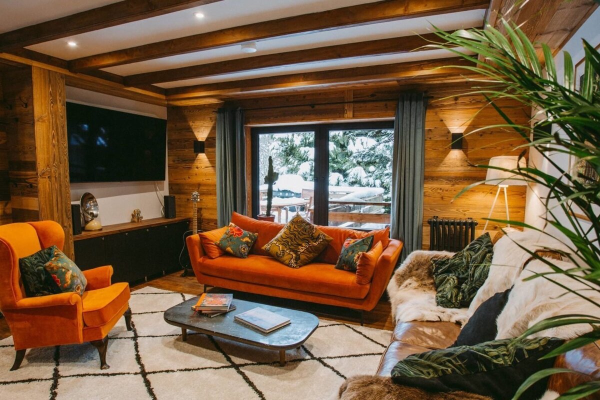 An inviting chalet living room featuring warm wood walls, a bright orange sofa, a modern TV, and a large window overlooking a snowy scene.