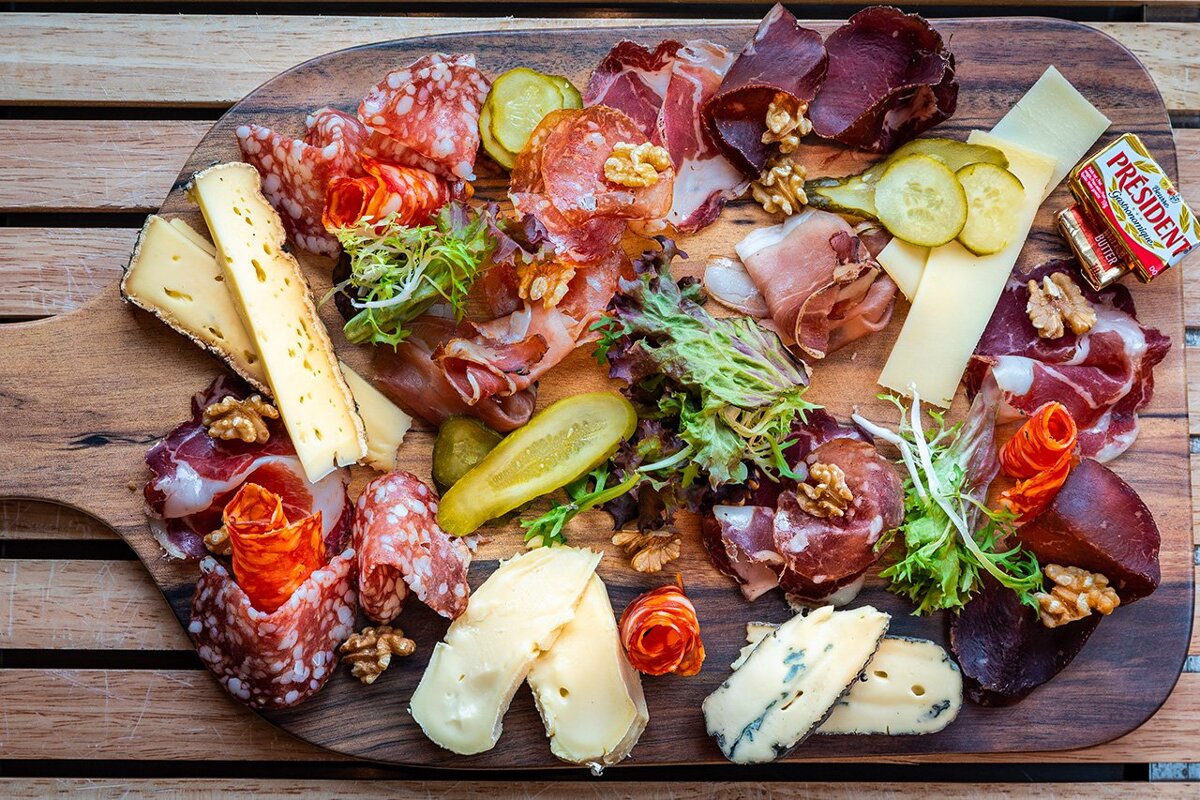 A delicious charcuterie and cheese board featuring sliced meats, various cheeses, pickles, walnuts, fresh greens, and a pat of butter.