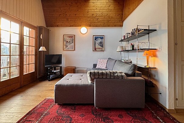 A rustic living room with a grey sectional, red oriental rug, wood-paneled ceiling, bright French doors, ski posters, TV, and modern shelving.