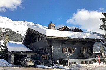 A snowy wooden chalet with cars parked, nestled among snow-covered mountains under a bright blue sky with sparse clouds.