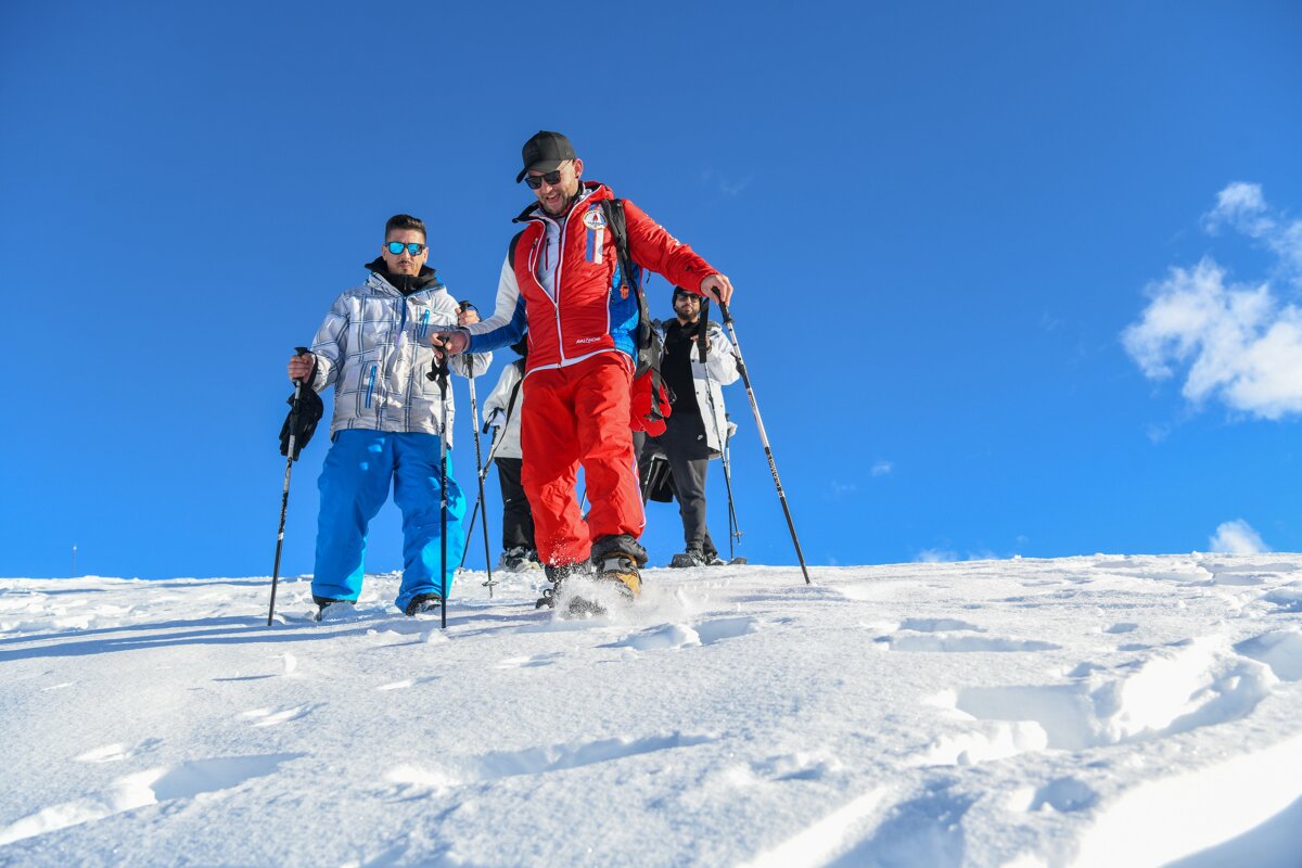 A group of people, equipped with poles and winter gear, snowshoe down a snowy slope under a vibrant blue sky.