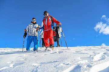 A group of people, equipped with poles and winter gear, snowshoe down a snowy slope under a vibrant blue sky.