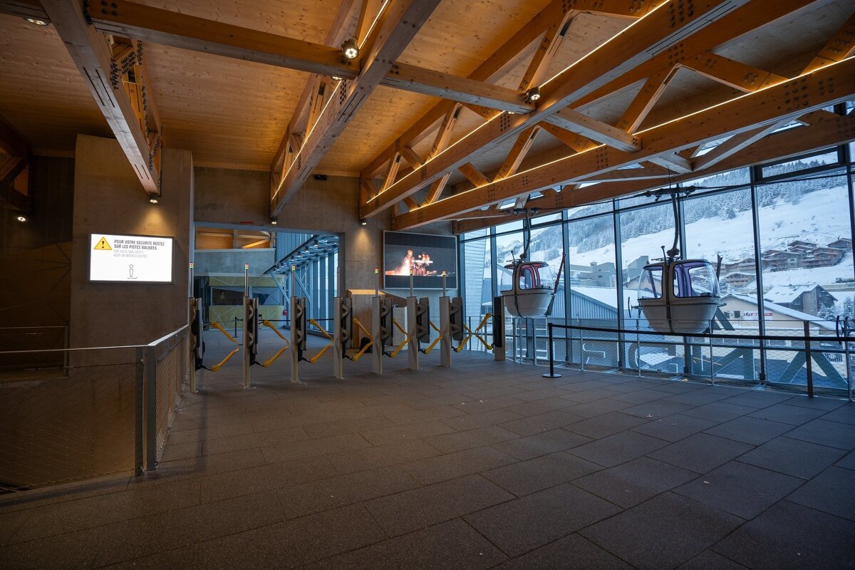 Modern gondola lift station interior: wooden ceiling, turnstiles, and cabins by a window revealing a snowy mountain village.