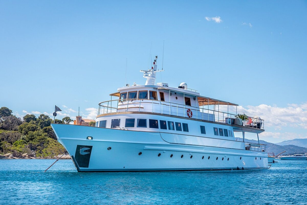 A white yacht floats in clear blue water, anchored near a green, tree-lined coast under a bright blue sky with light clouds.