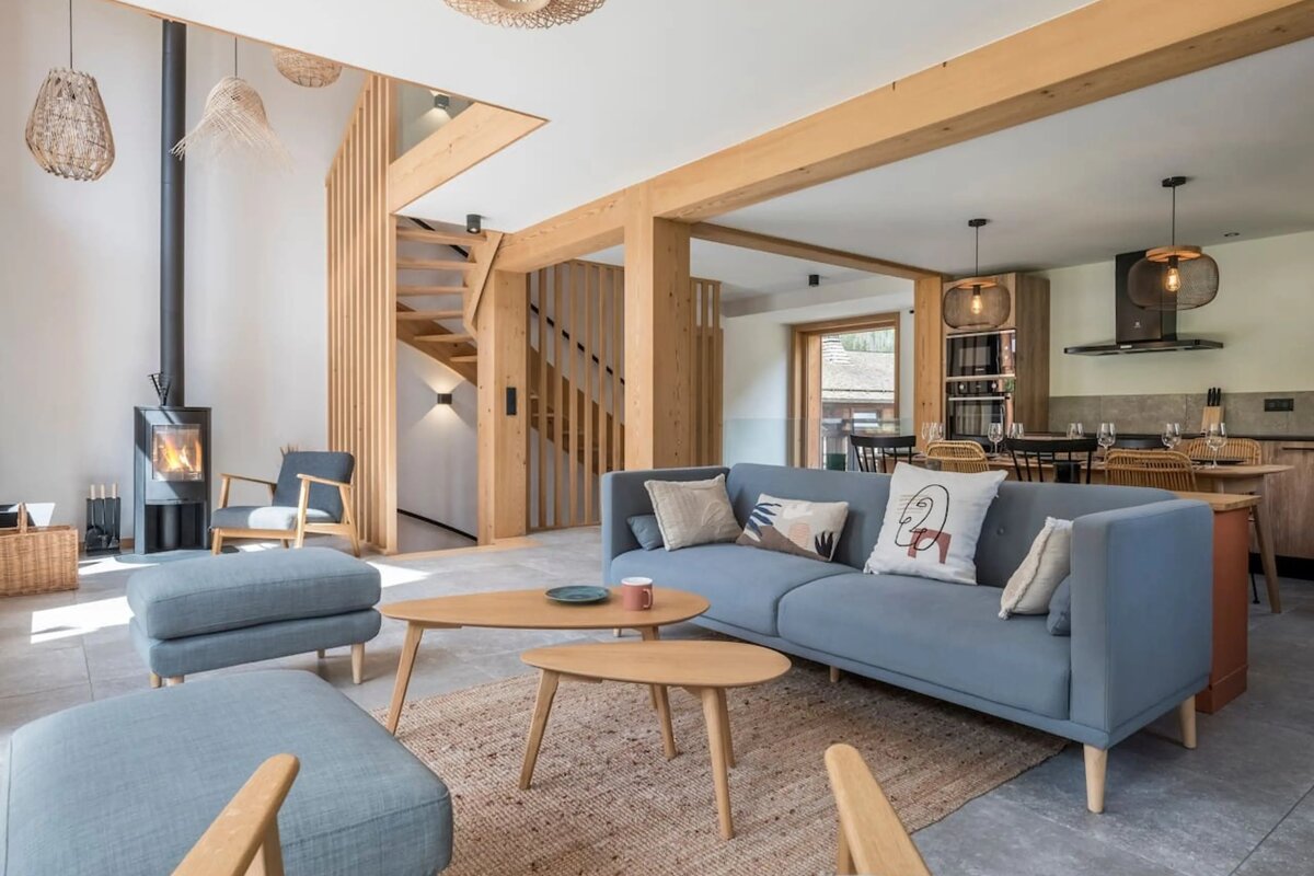 Modern open-plan living room featuring light wood accents, a blue sofa, wooden coffee tables, and a stylish staircase.