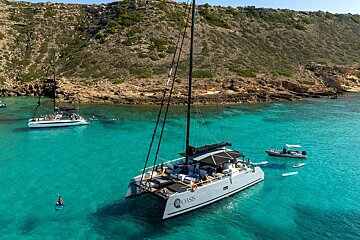 Two catamarans anchored in stunning turquoise water beside a rocky, green island. People are swimming, paddleboarding, and relaxing on deck.