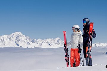 Two skiers stand on a snowy peak, looking out at a stunning view of snow-capped mountains above a sea of clouds.
