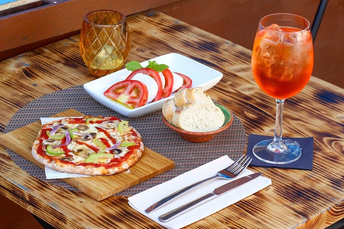 A casual meal on a rustic wooden table with a small pizza, Caprese salad, bread, and an orange iced drink.