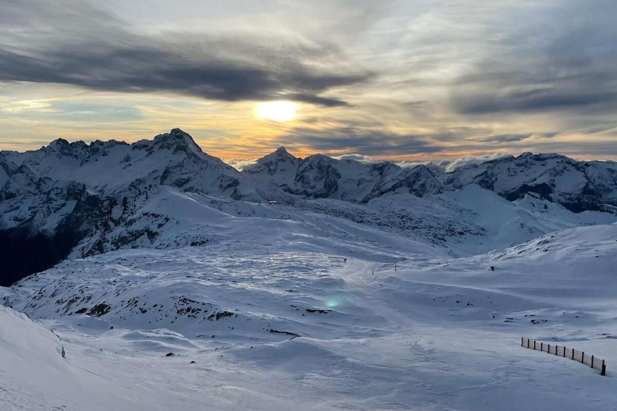 A golden sunset casts warm light over vast snow-covered mountains and wide slopes, under a cloudy sky, creating a serene winter landscape.