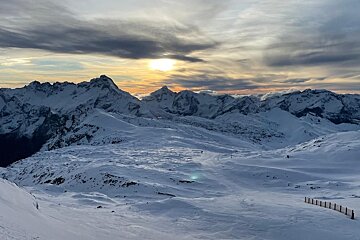 A golden sunset casts warm light over vast snow-covered mountains and wide slopes, under a cloudy sky, creating a serene winter landscape.