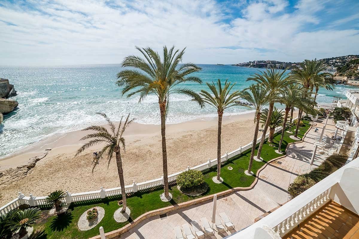 Tropical beach with tall palm trees lining a sandy shore, turquoise ocean waves, and a white resort building under a bright blue sky.