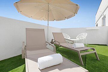 Sunny rooftop terrace with two loungers, an umbrella, and artificial grass, set against white walls and a clear blue sky.