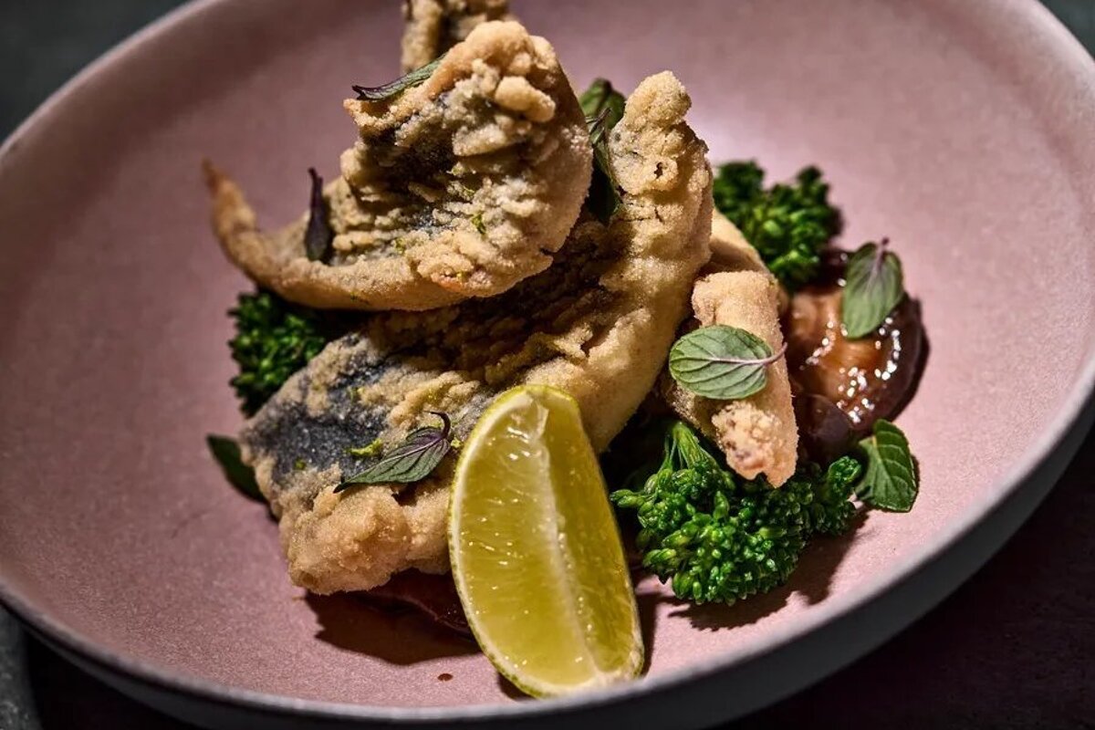 Crispy fried fish fillets, green broccolini, dark sauce, lime, and mint garnish in a stylish pink bowl.