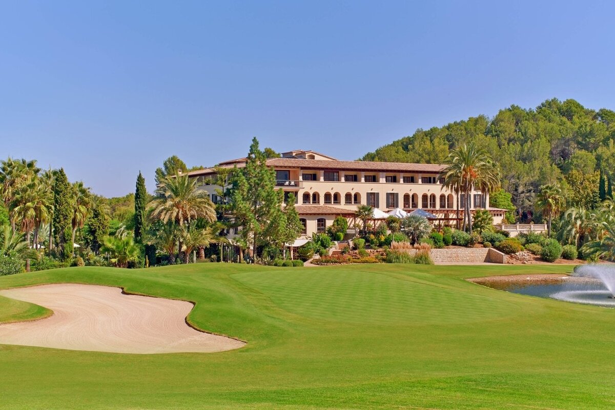 A grand hotel overlooks a lush golf course with a sand trap, palm trees, and a water fountain under a clear blue sky.