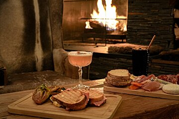 A cozy evening scene with food boards (grilled sandwiches, charcuterie) and drinks (pink cocktail, dark drink) on a wooden table by a warm fireplace.