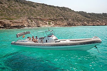 A white rigid inflatable boat with passengers floats on crystal-clear turquoise water near a rocky coastline.