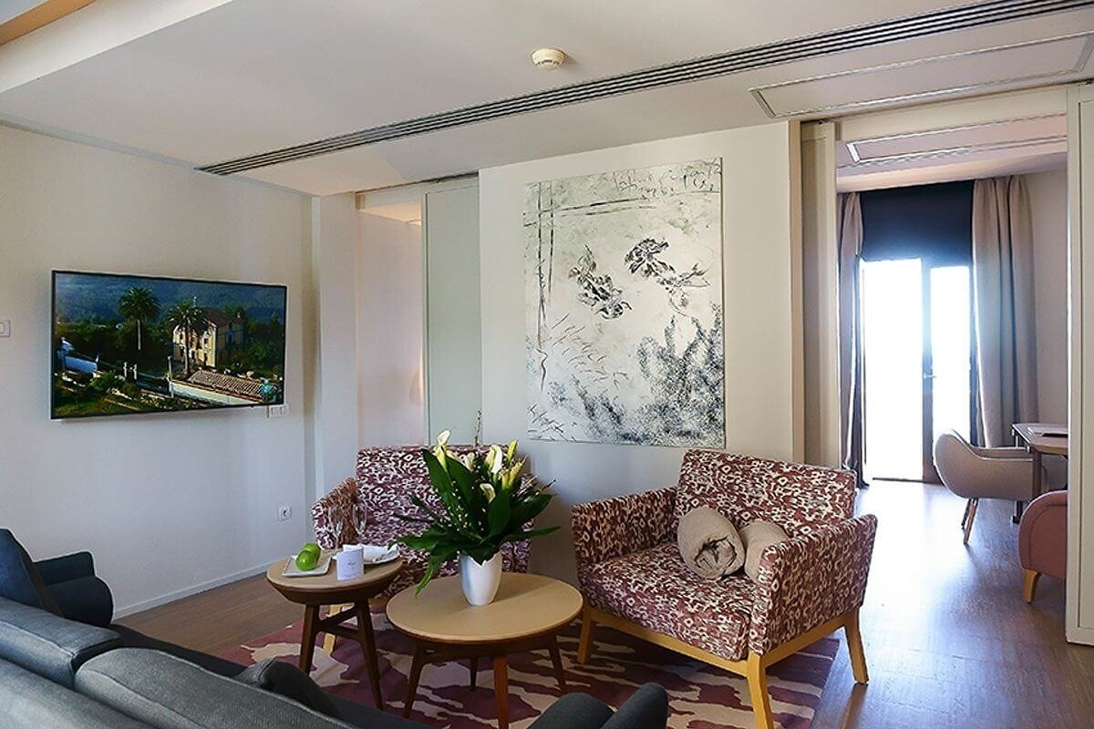 A modern, bright living room features a large TV, abstract wall art, two patterned armchairs, coffee tables, and a sunlit doorway.