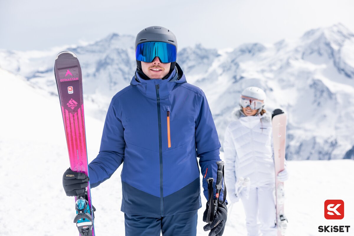 Two smiling skiers, a man in blue and a woman in white, stand with their skis and gear on a snowy mountain with majestic peaks in the background.