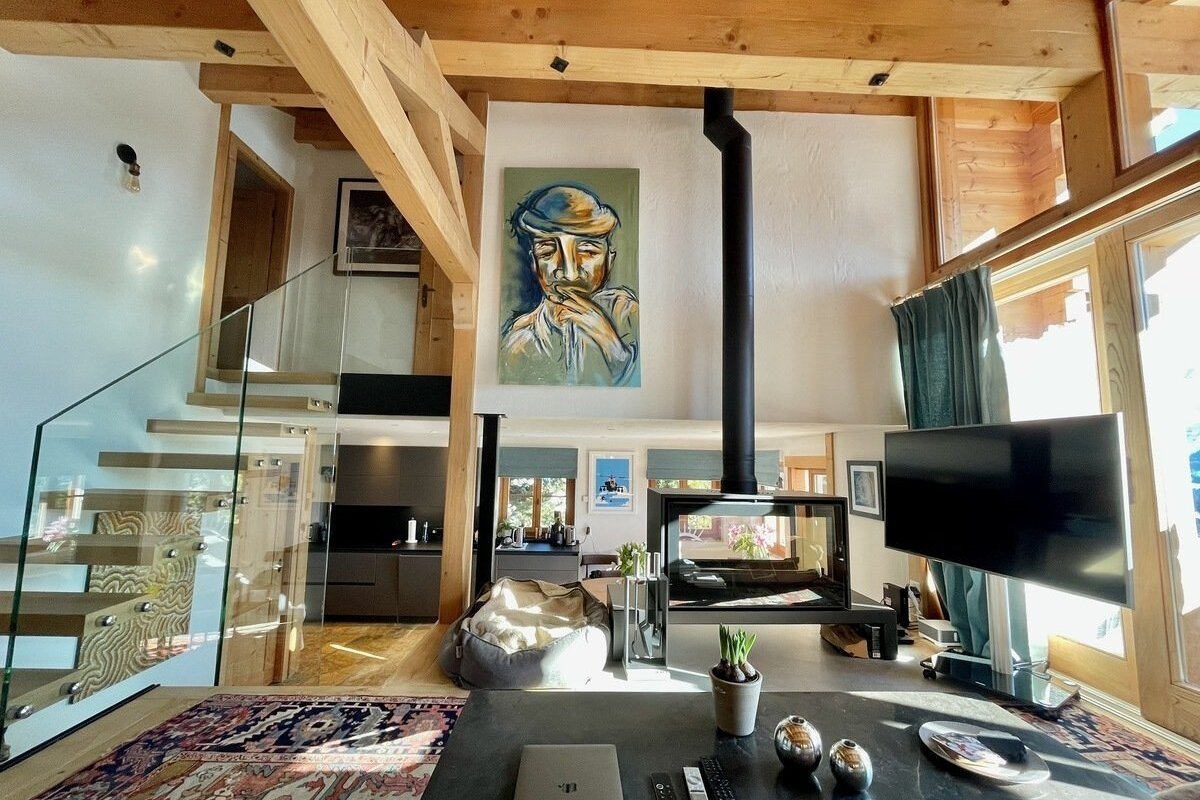 Bright, stylish open-plan living space with exposed wooden beams, a sleek glass staircase, modern suspended fireplace, and a large abstract portrait painting.